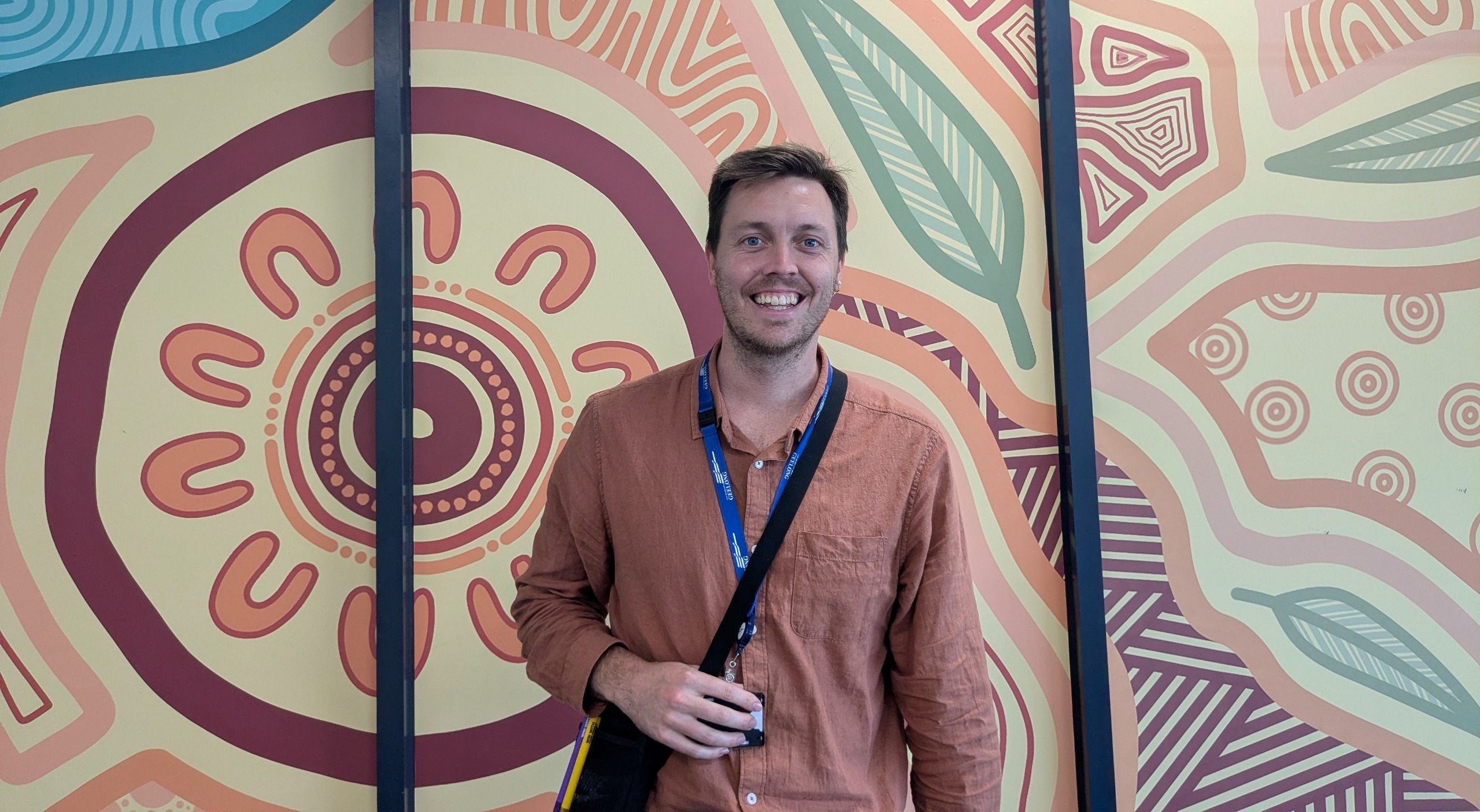 Ben is standing in front of a colourful First Nations mural. He is wearing a terracotta-coloured shirt and a blue lanyard, and is smiling at the camera. 
